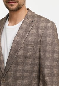 Brauner karierter Blazer aus strukturiertem Stoff mit Reverskragen, einem Knopfverschluss und einer Brusttasche. Trägt man über einem weißen T-Shirt.