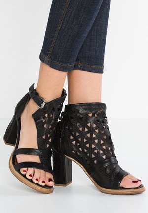 Sandales noires à talons hauts avec un design découpé géométrique, bride à boucle à la cheville, portées avec un jean court bleu foncé et des ongles d’orteils peints en rouge foncé.