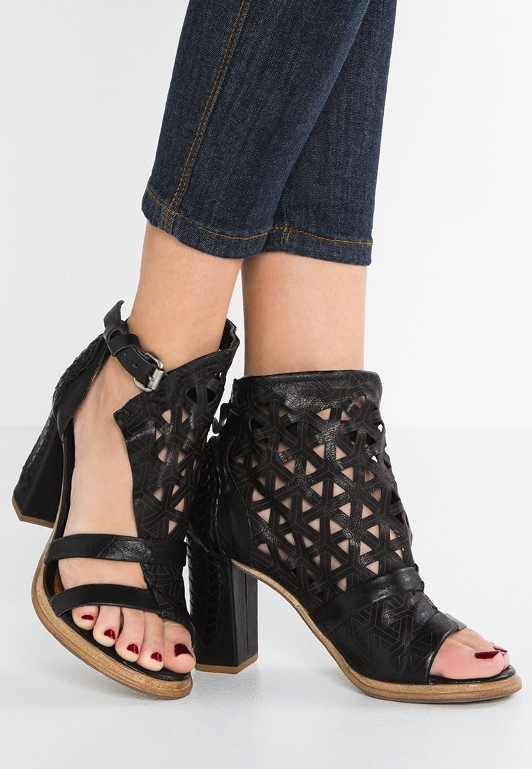 Sandales noires à talons hauts avec un design découpé géométrique, bride à boucle à la cheville, portées avec un jean court bleu foncé et des ongles d’orteils peints en rouge foncé.