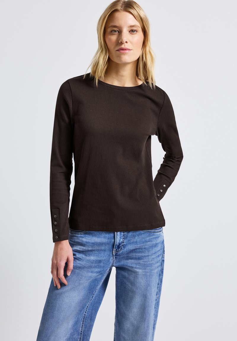 Rippstrick-Longsleeve-Shirt in Dunkelbraun mit rundem Ausschnitt und Knopfakzenten an den Ärmeln. Kombiniert mit hellblauen weit geschnittenen Jeans.