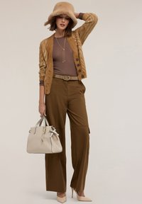 Nainen ruskeassa villapaidassa, leveälahkisissa housuissa, beigessä korkokenkäparissa ja hatussa pitää beigen käsilaukun, poseeraten neutraalia taustaa vasten.