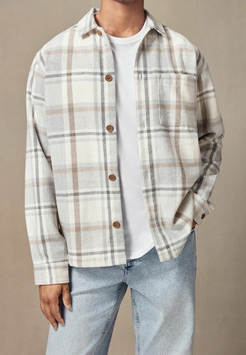 Homme portant une chemise à carreaux beige et grise boutonnée sur un t-shirt blanc, associée à un jean bleu clair, debout devant un fond neutre.