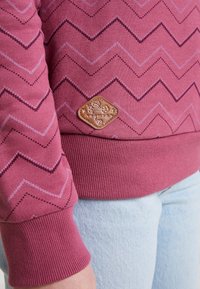 Primer plano de una persona que lleva un suéter de punto en patrón de chevron rosa con un parche de cuero marrón y jeans de color azul claro.