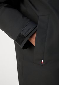 Svart vattentät jacka med en strukturerad yta, kardborreförslutning vid ärmslut och en logotypapplikation. Tydlig liten flaggadetail vid sidsömmen.