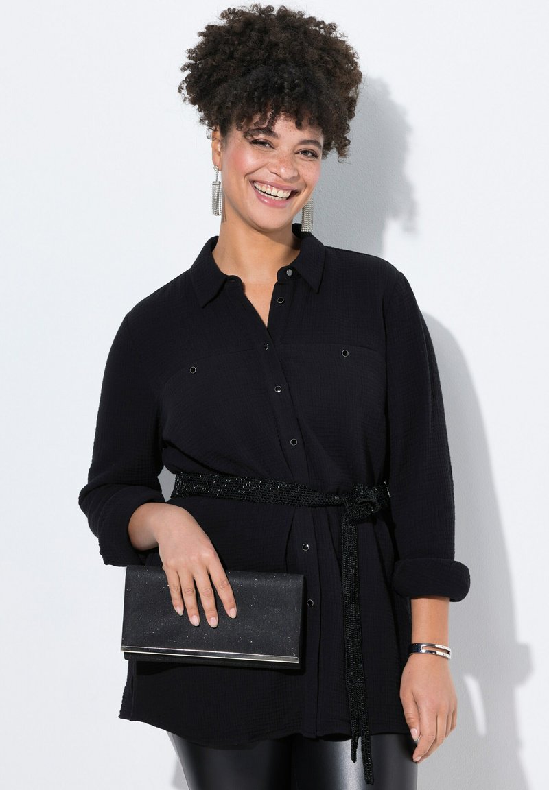 Schwarzes Hemd mit langen Ärmeln, strukturierter Stoff, tailliert mit einem glitzernden Gürtel; hält eine Clutch mit einem glänzenden Detail.