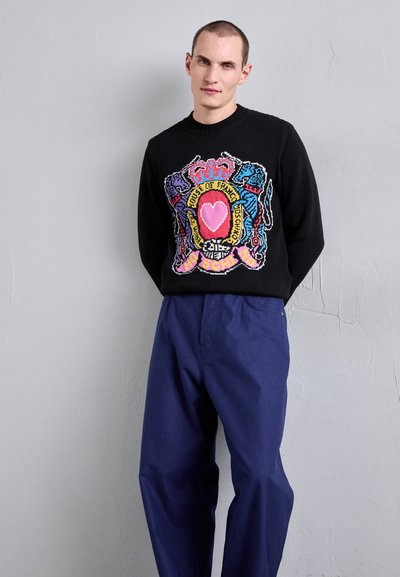 Svart stickad tröja med en färgglad crestmönstring med ett hjärta, kombinerad med löst sittande blå byxor. Enkla texturerad bakgrund.
