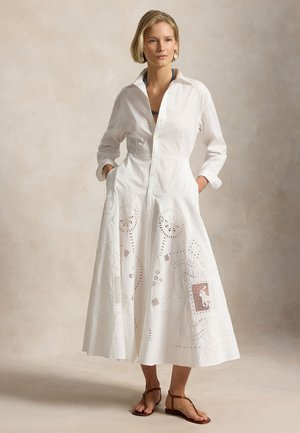 Kobieta w białej sukience z długim rękawem zapinanej na guziki, z haftowanymi wycięciami, stojąca z rękami w kieszeniach i ubraną w brązowe sandały.