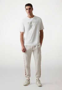 Fehér, környakú póló halvány háromszög logóval, krémszínű, oldalsó márkajelzéssel és rugalmas mandzsettával ellátott melegítőnadrággal, fehér tornacipővel kiegészítve.