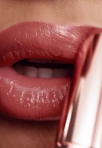 Gros plan sur des lèvres pulpeuses avec un rouge à lèvres rose clair et brillant. Le tube de rouge à lèvres a un boîtier métallique rose gold, mettant en valeur la texture lisse des lèvres.