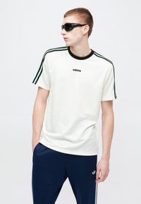 Fehér pamut póló fekete bordázott gallérral, zöld és fekete csíkokkal az ujjakon, valamint a mellkas közepén egy adidas logóval.