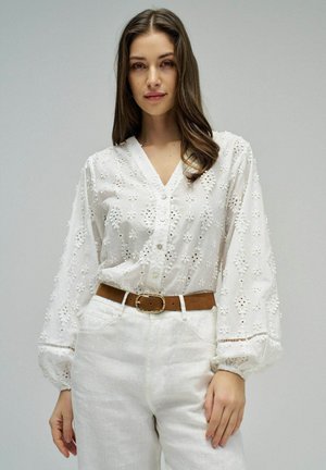 Mujer de cabello largo y castaño, vestida con una blusa blanca bordada con mangas abullonadas, pantalones blancos y un cinturón marrón, posando frente a un fondo gris.