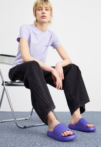 Lila gummislidesandaler med strukturerade band, bärs med svarta vida jeans. En minimalistisk, modern design på en ljus bakgrund.