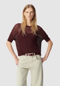 Bordeaux gebreide trui met diamantpatroon, korte mouwen, geribbelde halslijn, gedragen over lichtgekleurde wijde broek met een bijpassende riem.