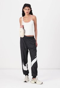 Calças desportivas pretas com um design de swoosh branco, combinadas com uma t-shirt sem mangas branca e uma bolsa transversal creme. Ténis completam o visual.