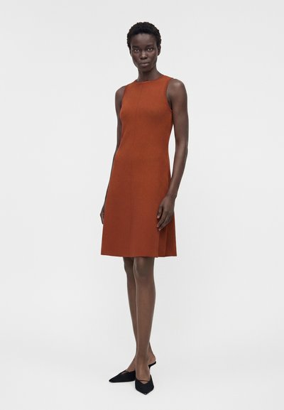 Γυναίκα στέκεται φορώντας ένα μανίκι χωρίς φόρεμα σε μήκος μέχρι το γόνατο σε καμμένο πορτοκαλί χρώμα και μαύρα μυτερά φλατ παπούτσια, με φόντο απλό ανοιχτό γκρι.