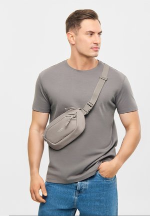 Bărbat purtând un tricou gri și blugi albaștri, cu o geantă gri purtată încrucișat pe piept, cu o curea reglabilă, uitându-se în lateral.