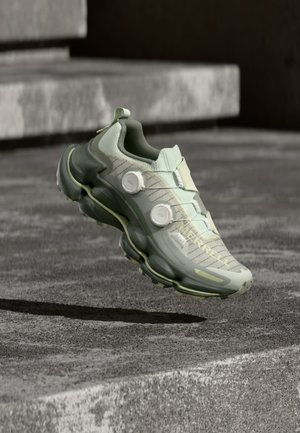 Hellgrüner und grauer Merrell-Sportschuh mit zwei Rädchen, schwebend über Betonstufen in einer Außenanlage.