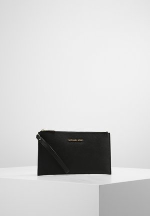 Pochette bracelet noire Michael Kors avec logo doré et fermeture éclair, exposée sur un socle blanc sur fond gris clair.
