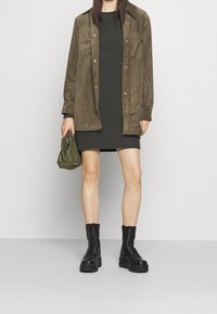Olive green corduroy shirt jacket with two chest pockets, worn over a dark gray ribbed dress. Black lace-up combat boots and an olive bag.