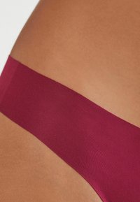 Burgundowy pas materiału bielizny, płaski i gładki w dotyku, kontrastujący z opaloną skórą. Zbliżenie, skupiające się na kolorze i materiale.
