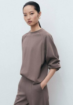 Bruine sweatshirt met een ontspannen pasvorm, raglanmouwen en een gestructureerde stof. Het model draagt ook bijpassende bruine broeken.