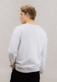 Hvit genser med ribbestrikkede ermer og nederkant, med en klassisk rund hals og en jevn tekstur, vist fra baksiden.