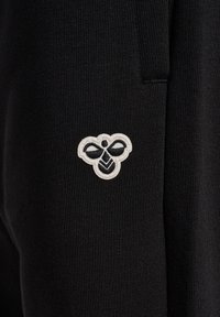 Schwarze Jogginghose mit gerippter Textur, die auf dem linken Bein ein kleines, weißes gesticktes Emblem in Form einer stilisierten Biene zeigt.