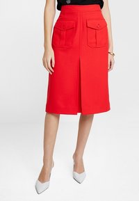 Falda roja hasta la rodilla de tela suave, con una abertura frontal y dos bolsillos con tapa con cierres de botón. Combinada con tacones gris claro.