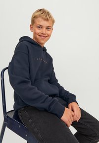 Felpa blu navy in pile spazzolato con tasca frontale e logo sul petto, abbinata a jeans neri. Seduto su una sedia.