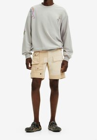 Sudadera gris claro con detalles gráficos, combinada con pantalones cortos tipo cargo beige que cuentan con múltiples bolsillos y herrajes negros. Zapatos negros sin cordones.