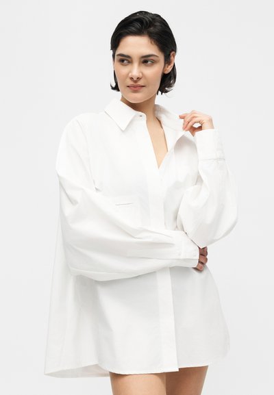 Γυναίκα με κοντά σκούρα μαλλιά που φοράει μια υπερμεγέθη λευκή πουκαμίσα με κουμπιά, κρατώντας το γιακά με το ένα χέρι, πάνω σε μια μονόχρωμη φόντο.