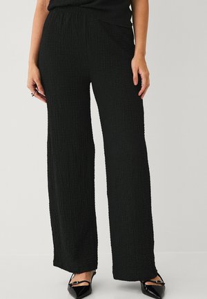 Kvinde iført højttaljede, vidde bukser i sort tekstureret stof, kombineret med sorte spidse sko med anklestropper.