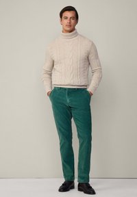 Pull en maille torsadée beige avec un col montant associé à un pantalon en velours côtelé vert et des mocassins noirs. Le mannequin se tient avec les mains dans les poches devant un fond neutre.