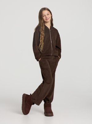 Set coordinato marrone in velluto a coste con felpa con cappuccio e pantaloni, chiusura frontale con zip. Presenta maniche larghe, tasche frontali e tessuto morbido al tatto. Stivaletti marroni alla caviglia.