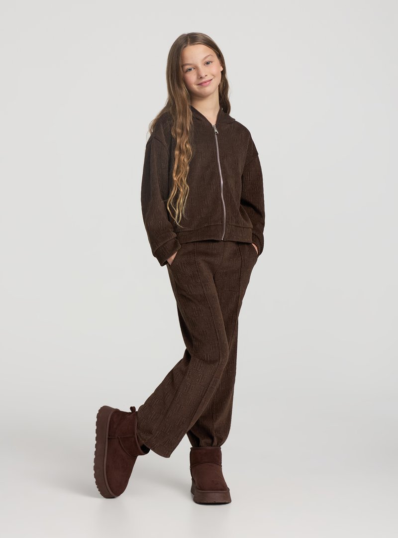 Set coordinato marrone in velluto a coste con felpa con cappuccio e pantaloni, chiusura frontale con zip. Presenta maniche larghe, tasche frontali e tessuto morbido al tatto. Stivaletti marroni alla caviglia.