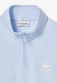 Pullover azzurro chiaro, con vestibilità classica e chiusura a zip, dotato di colletto. Presenta un tessuto testurizzato e un logo Lacoste bianco sul petto.