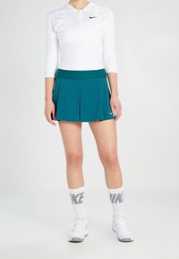 Camiseta atlética blanca de manga larga con media cremallera, falda plisada azul petróleo y zapatillas blancas con detalles negros, combinadas con calcetines hasta la rodilla.