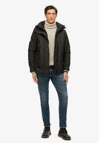 Zwarte pufferjack met een capuchon, gestructureerde grijze coltrui, blauwe jeans en zwarte veterschoenen, staand tegen een witte achtergrond.