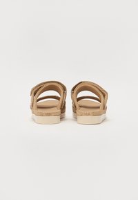 Tan semskinn sandaler med åpen bak, flere stropper og polstring. Har en logo på hælen og en lys gummisåle.