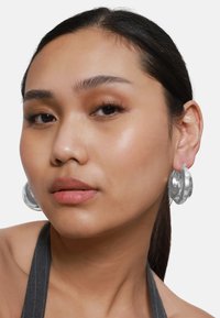 De grandes boucles d'oreilles en argent avec une texture lisse et réfléchissante, ornées d'un design en spirale. Portées par un modèle sur un fond neutre.