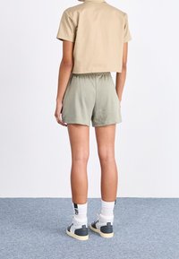 Béžové krátké tričko a olivově zelené šortky z lehké látky. Modelka má na sobě bílé a šedé tenisky s kontrastními detaily.