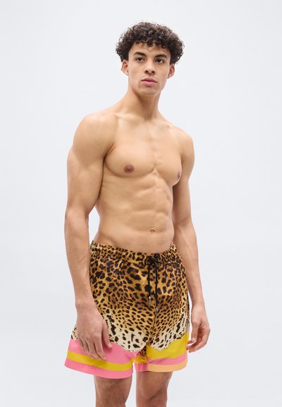 Άνδρας χωρίς μπλούζα με σγουρά μαλλιά, που φοράει μαγιό με λεοπάρ σχέδιο και ροζ και κίτρινες ρίγες, κοιτώντας προς το πλάι.