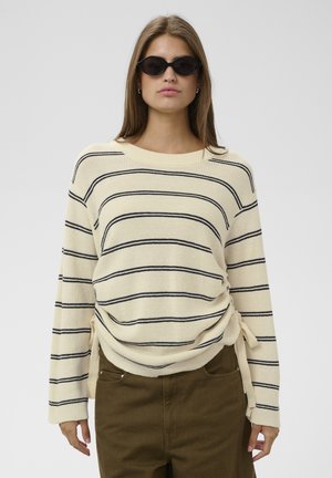 Femme portant des lunettes de soleil ovales noires, un pull crème avec des rayures horizontales noires et un pantalon marron, debout devant un fond clair uni.
