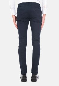 Pantaloni slim-fit di colore blu navy realizzati in un tessuto resistente. Presentano tasche posteriori con zip, passanti per cintura e una texture liscia. Scarpe nere visibili.