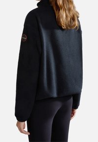 Pullover in felpa nera con colletto alto, superficie testurizzata e una toppa rotonda con logo sulla spalla sinistra; abbinato a leggings scuri.