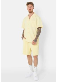 Camisa de manga corta con botones y pantalones cortos a juego en suave tela amarilla. Cuenta con un bolsillo en el pecho y un corte relajado, combinada con zapatillas blancas.