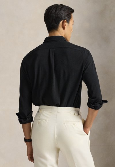 Vyras dėvi juodą ilgomis rankovėmis marškinius su susuktais rankovėmis ir baltas pritaikytas kelnės, stovintis su ranka kišenėje, atsuktas nugarą.