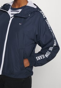 Navy blue zip-up jacket with hood, white trim, "Tommy Jeans" text on sleeve, small logo on chest, worn over striped shirt, hand in pocket.