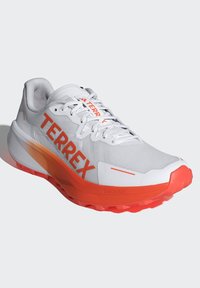Hvide løbesko med orange accenter og stort "TERREX" mærkning. Har en struktureret overdel, polstret krave og rillede såler for bedre greb.