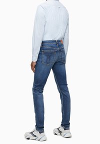 Modré džíny s úzkým střihem, s zadními kapsami s unikátními švy, kombinované se světle modrou košilí s dlouhým rukávem a teniskami.